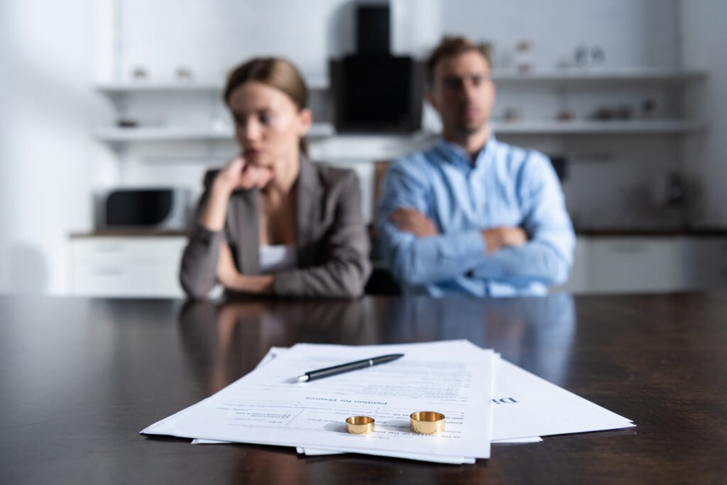Pareja sentada separada en una mesa con los papeles del divorcio y los anillos de boda en primer plano.
